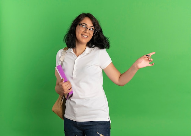 Молодая радостная симпатичная кавказская школьница в очках и задней сумке смотрит и указывает на бок, держа книги на зеленом с копией пространства