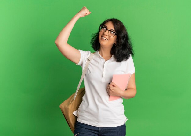 Young joyful pretty caucasian schoolgirl wearing glasses and back bag looks and raises fist up holding notebook on green  with copy space