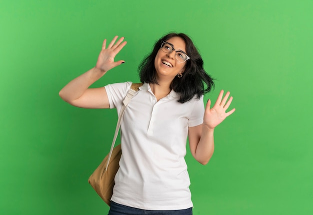 Young joyful pretty caucasian schoolgirl wearing glasses and back bag looks up with raised hands on green  with copy space