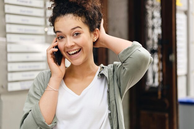 Young joyful woman with dark curly hair in khaki shirt and white T-shirt happily looking in camera and talking on cellphone on city street