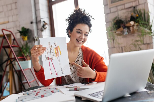 Young joyful woman with dark curly hair sitting at the table happily showing fashion illustrations in laptop spending time in modern cozy workshop with big windows
