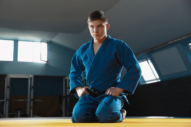 Young judo caucasian fighter in blue kimono with black belt posing confident in the gym, strong and healthy.