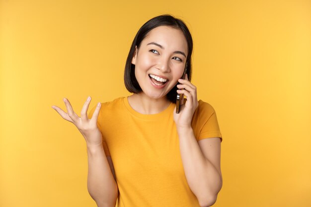 携帯電話で話している若い韓国の女の子黄色の背景の上に立っているスマートフォンで呼び出すアジアの女性