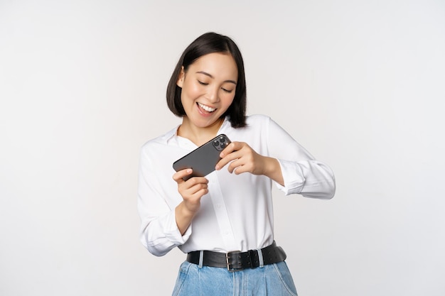 Young korean woman asian girl playing mobile video game on smartphone looking at horizontal phone screen standing over white background