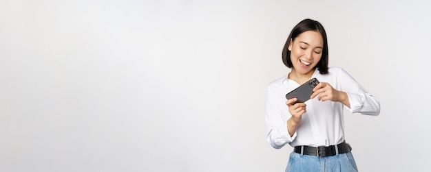Free photo young korean woman asian girl playing mobile video game on smartphone looking at horizontal phone screen standing over white background