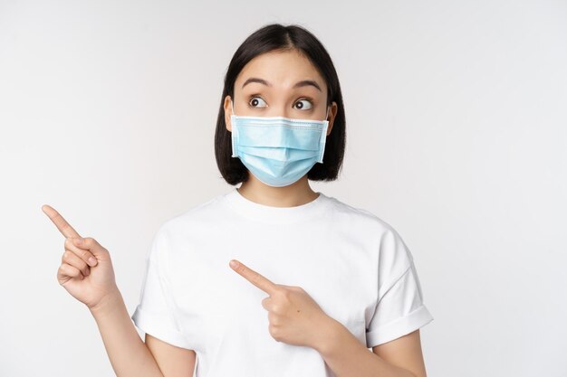 Young korean woman in medical face mask pointing fingers left and looking at logo showing advertisement or banner standing over white background