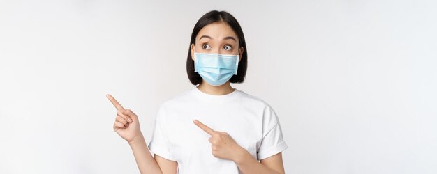 의료용 얼굴 마스크를 쓴 젊은 한국 여성이 손가락을 왼쪽으로 가리키고 흰색 배경 위에 서 있는 광고 또는 배너가 표시된 로고를 보고 있습니다.