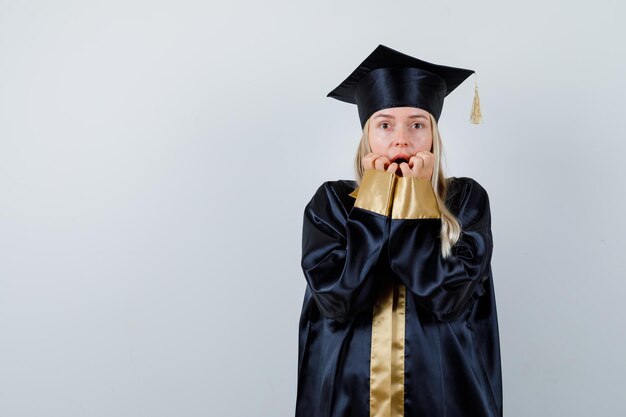 Young lady in academic dress keeping hands on mouth and looking scared