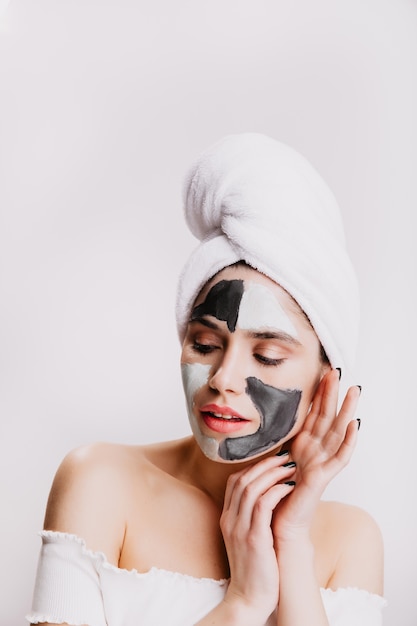 Young lady after shower is enjoying spa facial procedure. Portrait of woman in clay mask on white wall.