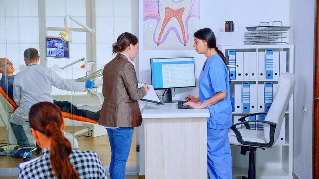 Young lady asking informations filling in stomatological form while patients talking sitting on chair in waiting area. People speaking in crowded professional orthodontist reception office.