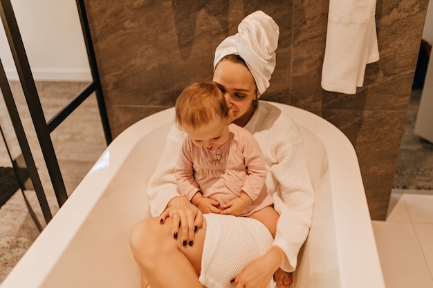Young lady in bathrobe and towel lying in bathroom. Cute child sits on mom and plays.