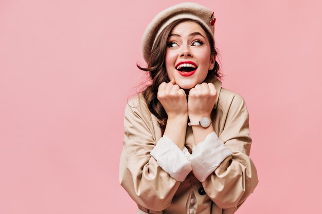 Young lady in beige jacket and beret dreamily posing on pink background.