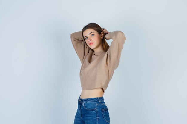 Young lady in beige sweater, jeans posing while with hands behind head and looking confident , front view.