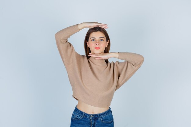 Молодая дама в бежевом свитере, джинсах показывает знак размера и выглядит уверенно, вид спереди.