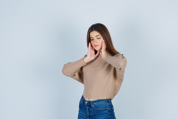 Young lady in beige sweater, jeans with hands on cheeks and looking confident , front view.