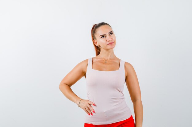 Young lady in beige tank top holding hand on waist and looking thoughtful , front view.