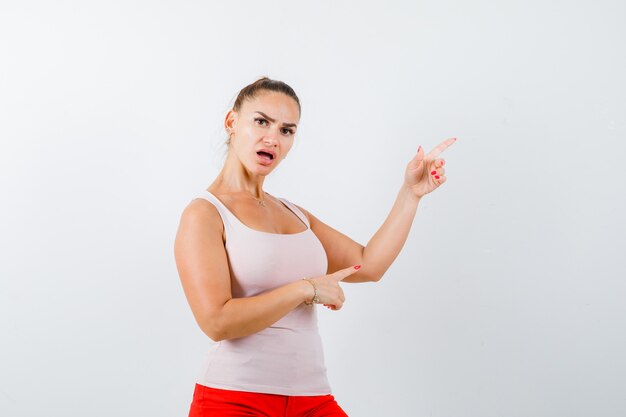 Young lady in beige tank top pointing aside and looking anxious , front view.