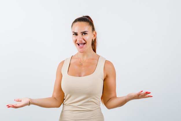 Young lady in beige tank top spreading palms aside and looking happy , front view.