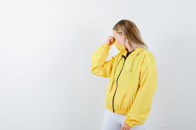 Young lady bending head down in jacket, pants, mask and looking depressed , front view.