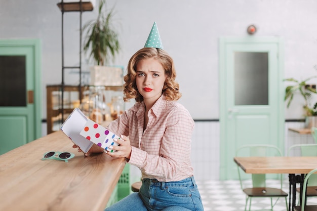 Young lady in birthday cap sitting at the bar counter with open present box in hands and sadly looking in camera in cafe