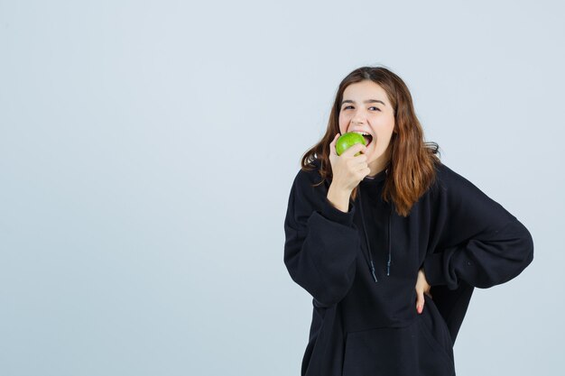 Юная леди кусает яблоко, держа руку на талии в огромной толстовке с капюшоном, штанах и выглядит довольной. передний план.