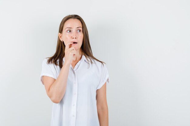 Young lady biting her finger in white blouse and looking thoughtful.