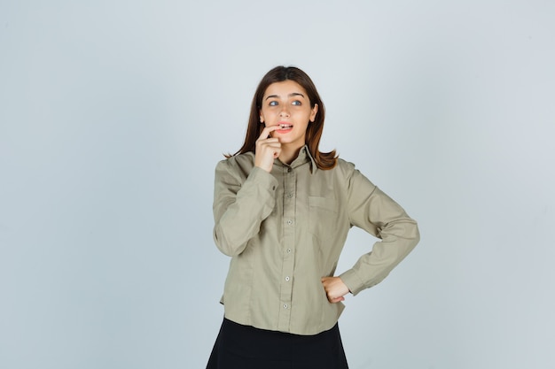 Young lady biting her nails in shirt, skirt and looking forgetful