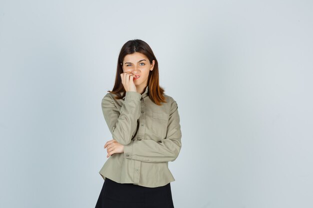 Young lady biting her nails in shirt, skirt and looking thoughtful