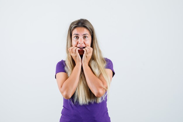 Young lady biting nails in violet t-shirt and looking scared , front view.