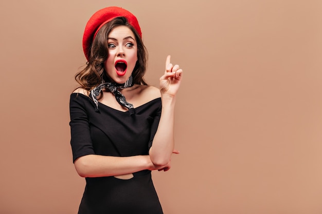 Девушка в черном платье, красной шляпе и шарфе придумала новую идею и показывает указательным пальцем вверх.