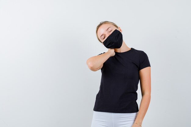 검은 색 티셔츠에 젊은 아가씨, 목 통증으로 고통 받고 피곤해 보이는 마스크, 전면보기.