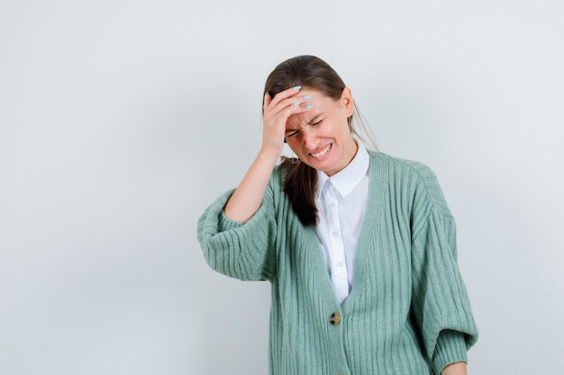 Young lady in blouse, cardigan with hand on forehead while crying and looking sorrowful , front view.