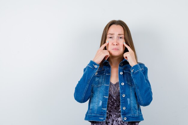 Young lady in blouse, denim jacket holding fingers on cheeks while crying and looking offended , front view.