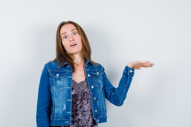 Young lady in blouse, denim jacket holding something and looking puzzled , front view.