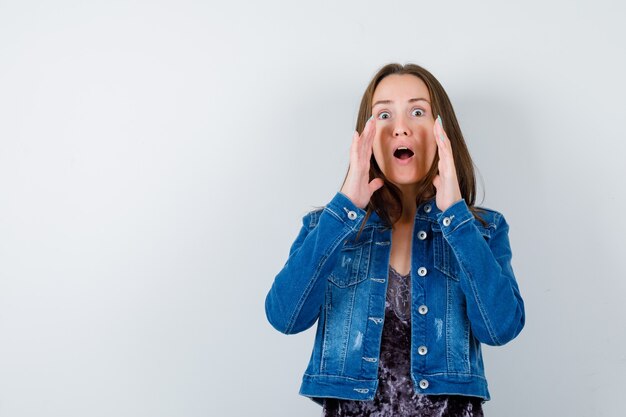 Young lady in blouse, denim jacket shouting something with hands and looking puzzled , front view.