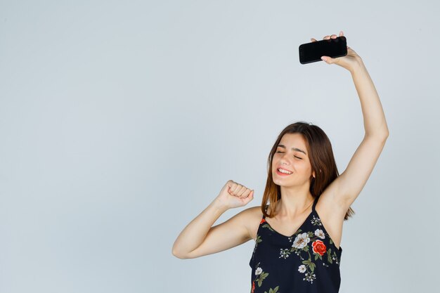 Young lady in blouse holding smartphone, showing winner gesture and looking happy , front view.