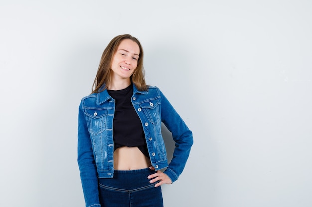 Young lady in blouse, jacket, jeans posing with hand on waist and looking confident, front view.