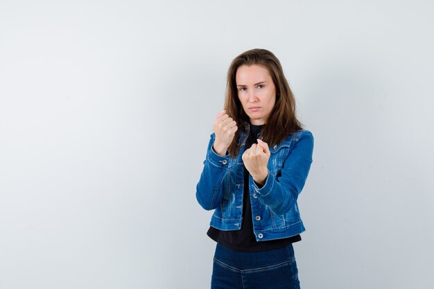 Young lady in blouse, jacket, jeans standing in fight pose and looking confident, front view.