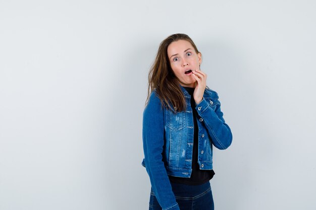 Young lady in blouse jeans holding hand near open mouth and looking surprised , front view.