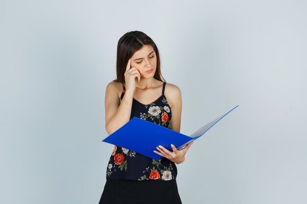 Young lady in blouse looking over folder, holding finger on temples and looking thoughtful , front view.