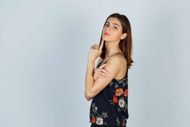 Young lady in blouse posing while touching her arm and chin and looking graceful .