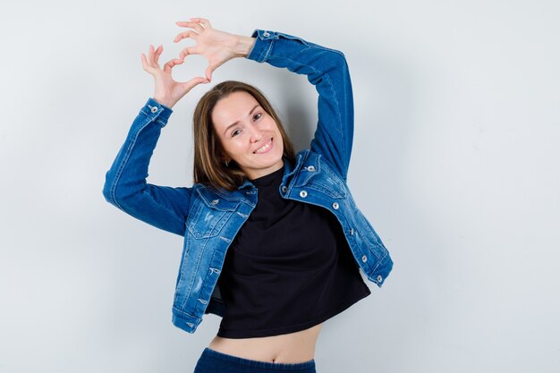 Young lady in blouse showing heart gesture and looking merry, front view.