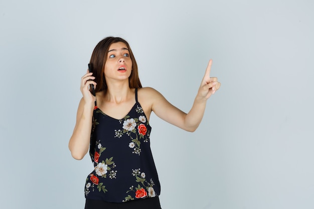 Free photo young lady in blouse showing hold on a minute gesture while talking on smartphone and looking careful , front view.