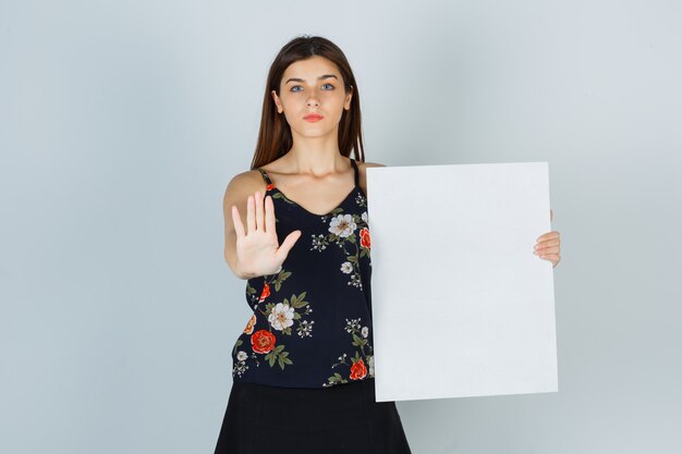 Young lady in blouse, skirt holding blank canvas, showing stop gesture and looking serious , front view.