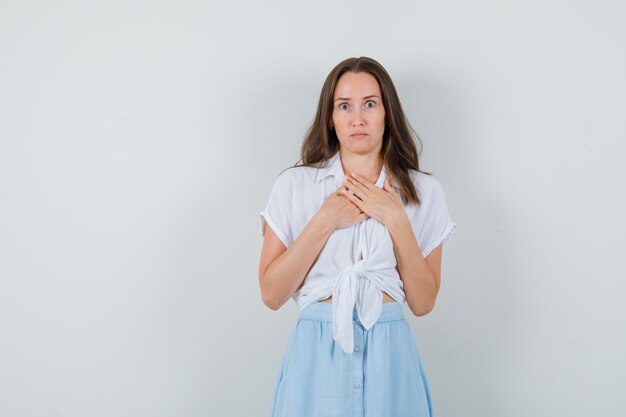 Young lady in blouse, skirt holding hands on her chest and looking worried