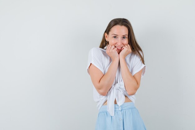 Young lady in blouse, skirt holding hands on her chin while smiling and looking glad