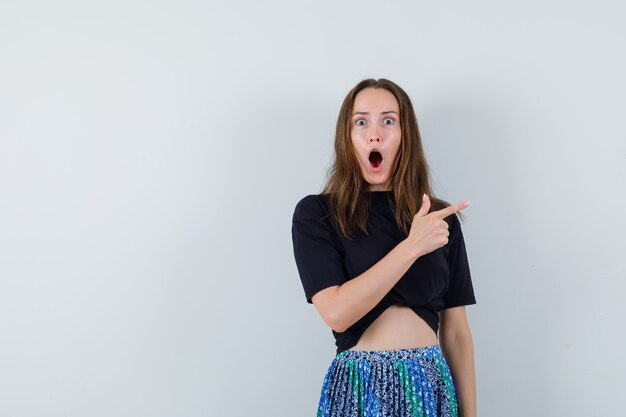 Young lady in blouse, skirt pointing aside and looking surprised