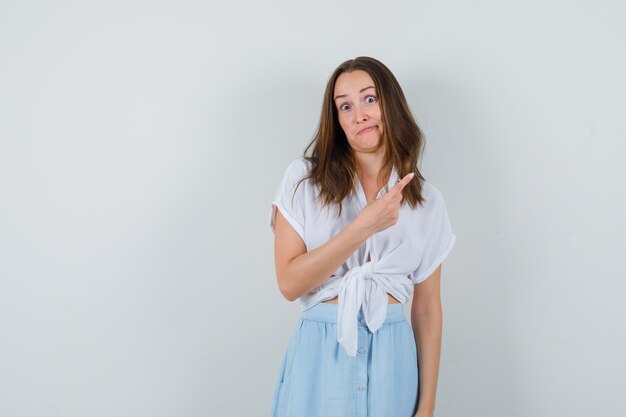 Young lady in blouse and skirt pointing at upper right corner and looking confused