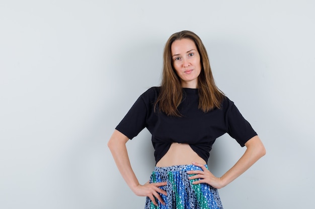 Young lady in blouse, skirt posing with hands on waist and looking cute