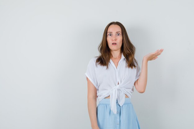 Young lady in blouse, skirt raising open palm and looking perplexed